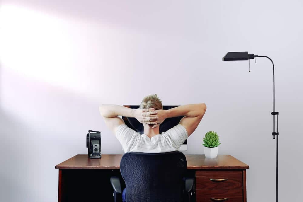 photograph of a man seen from behind in front of his laptop with his hands behind his head to illustrate online meeting exhaustion