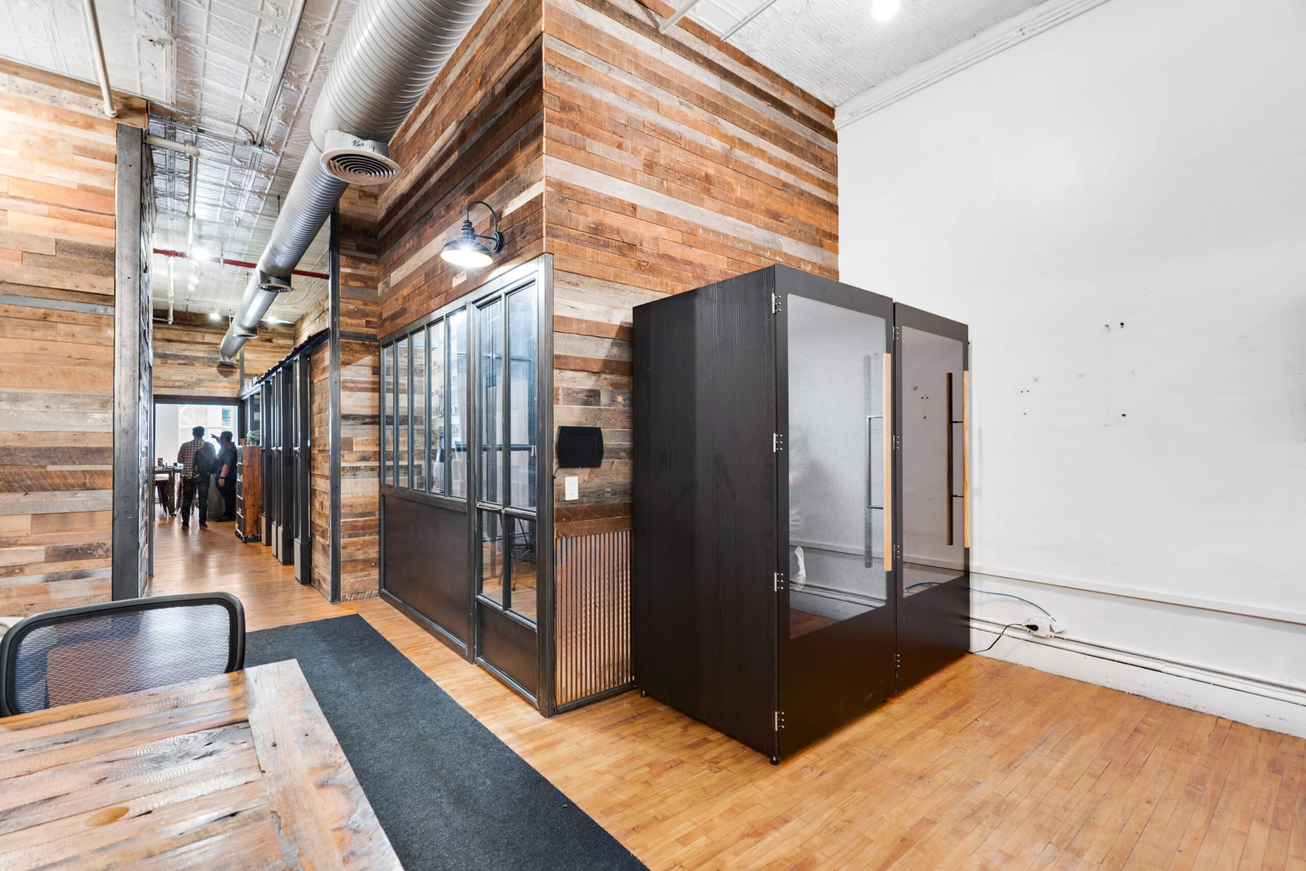 An office environment showcasing wooden walls and a wooden table, alongside phone booths and a communal workspace.